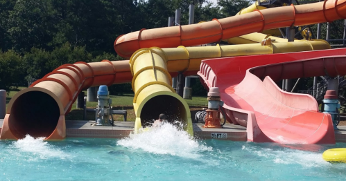 Colorful water slides at a pool, with splashes as riders enter the water below. Green trees in the background.