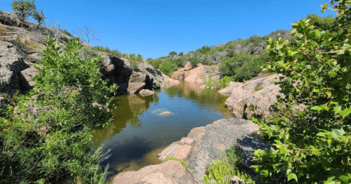 A serene landscape featuring a calm pond surrounded by rocky terrain and lush greenery under a clear blue sky.