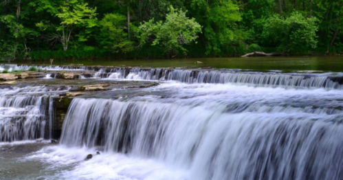 A serene waterfall cascades over rocky ledges, surrounded by lush green trees and a calm river.