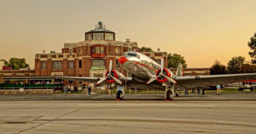 A vintage airplane parked in front of a historic airport terminal at sunset.