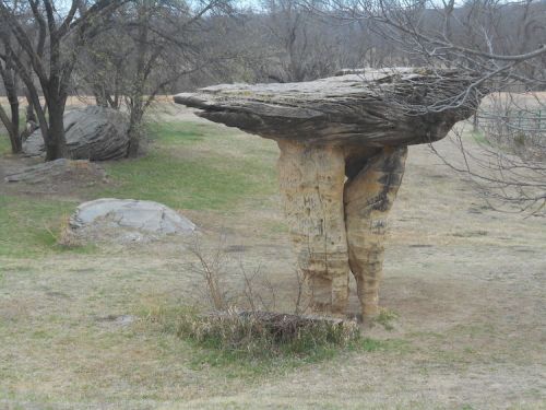 A large, flat-topped rock formation stands on a grassy area, surrounded by trees and smaller rocks.