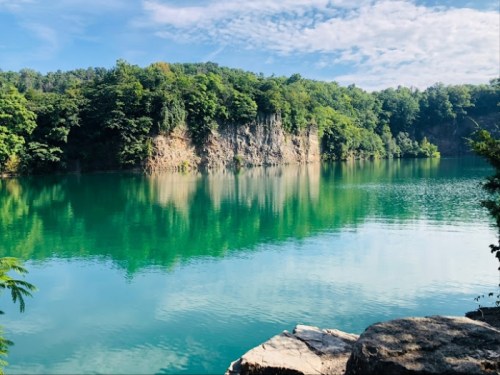 A serene lake surrounded by lush greenery and rocky cliffs, reflecting the blue sky and clouds above.