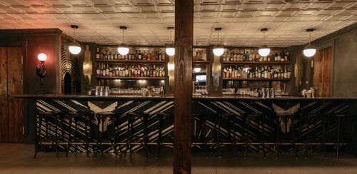A stylish bar with a dark wood counter, shelves of liquor, and vintage lighting in a cozy, modern setting.