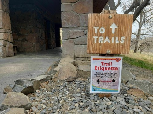 Wooden sign pointing to trails with a poster about trail etiquette, placed near a stone building and gravel path.