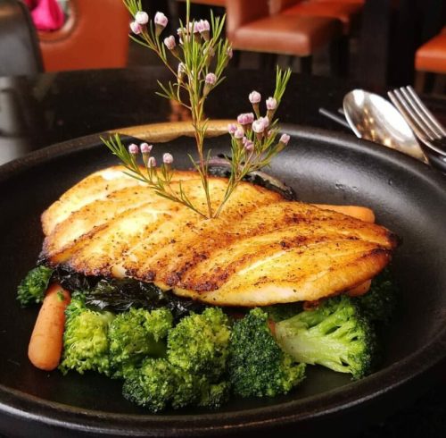 A beautifully plated dish featuring grilled fish atop broccoli and carrots, garnished with a small flower.