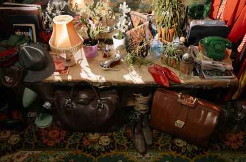 A cluttered table with various vintage items, including bags, a lamp, plants, and books, set against a colorful background.