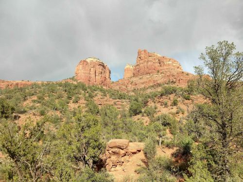 Red rock formations rise above a green landscape under a cloudy sky, showcasing the natural beauty of the area.