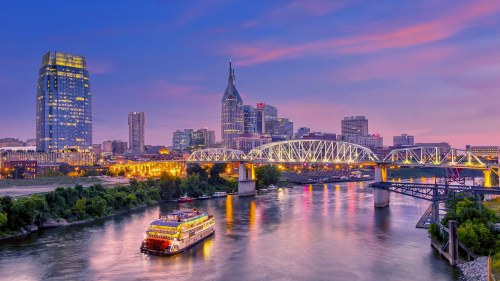 A scenic view of a city skyline at sunset, featuring a riverboat on the water and illuminated buildings and bridges.