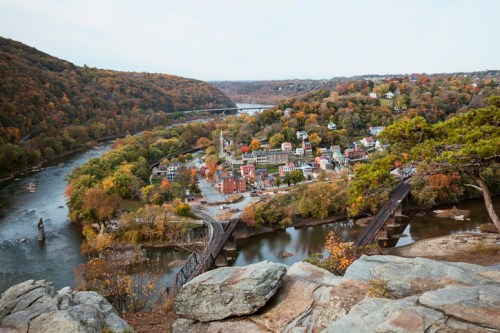A scenic view of a river town surrounded by autumn foliage, with a bridge and hills in the background.