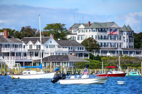 A scenic waterfront view featuring boats and historic houses with American flags, under a clear blue sky.