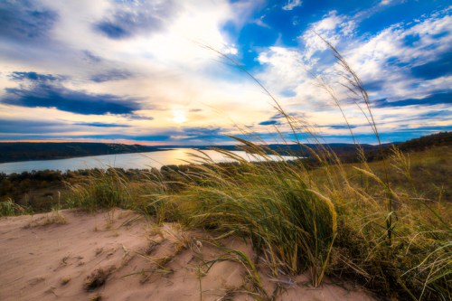 A serene landscape featuring tall grass in the foreground, with a lake and dramatic clouds at sunset in the background.