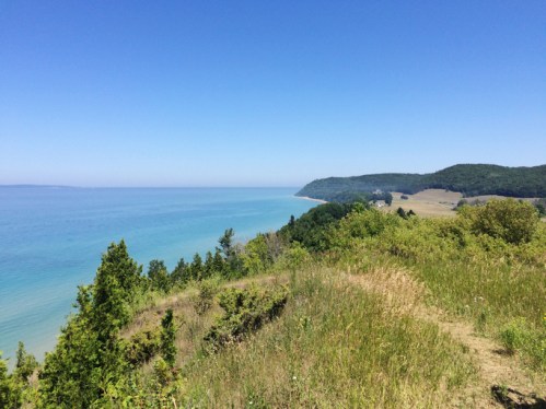 A scenic view of a calm blue lake bordered by green hills and clear skies on a sunny day.