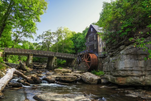 A scenic view of a historic watermill beside a flowing stream, surrounded by lush greenery and a wooden bridge.