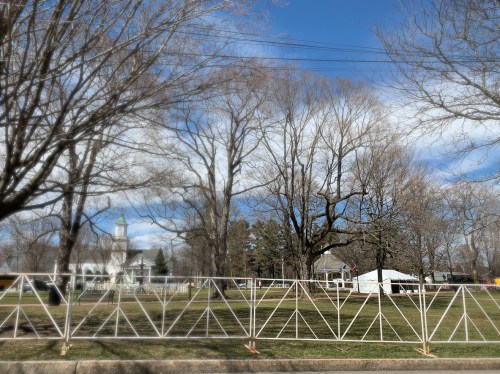 A park scene with bare trees, a church in the background, and a white tent set up on a grassy area under a blue sky.