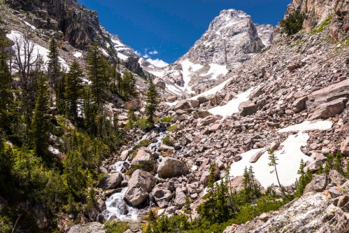A rocky mountain landscape with a stream, snow patches, and evergreen trees under a clear blue sky.