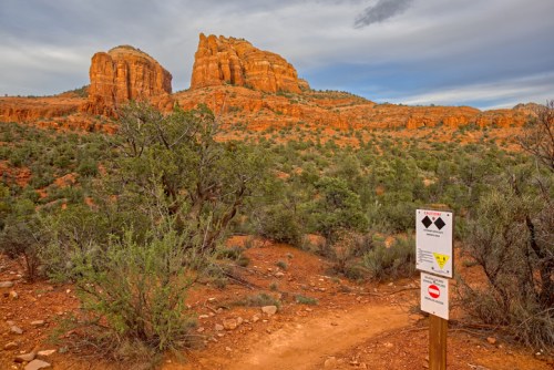 A desert landscape featuring red rock formations, green shrubs, and a trail sign indicating caution.