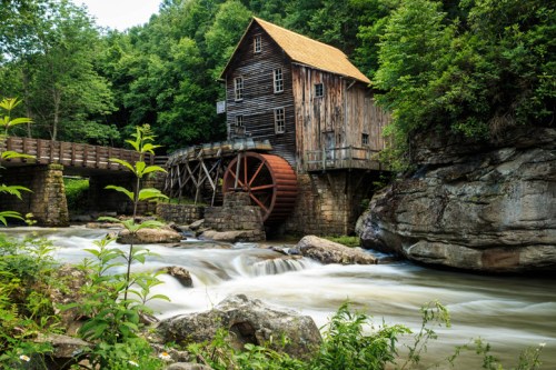 A rustic wooden mill with a water wheel beside a flowing river, surrounded by lush green trees and rocky terrain.