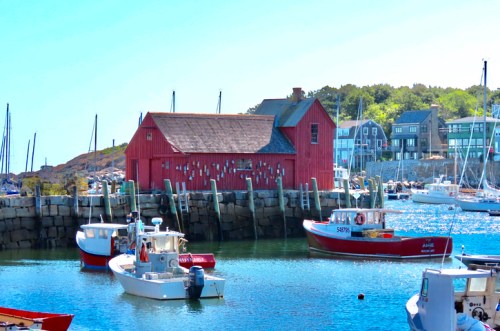 A scenic harbor with colorful boats and a red building, surrounded by calm water and green trees in the background.
