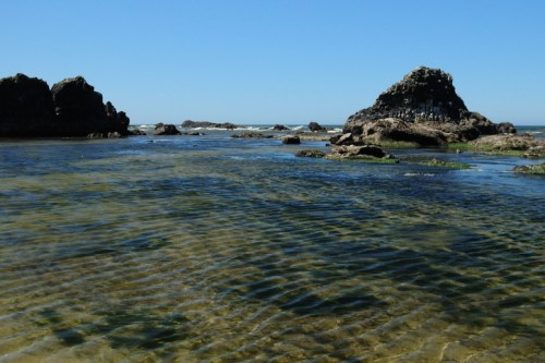 A serene coastal scene with clear water, rocky formations, and a bright blue sky.