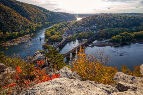 A scenic view of a river winding through hills, with a bridge and colorful autumn foliage in the foreground.