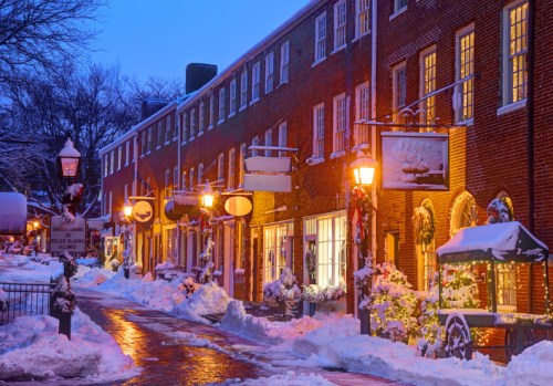 Snow-covered street lined with brick buildings, warm lights glowing, and festive decorations in a winter evening scene.