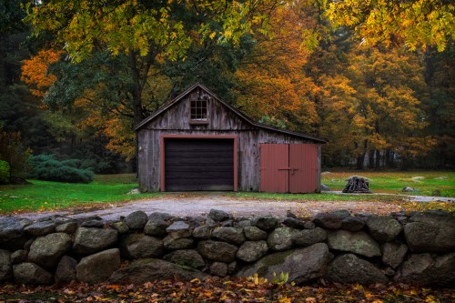 A rustic wooden barn with a red door, surrounded by colorful autumn foliage and a stone wall.