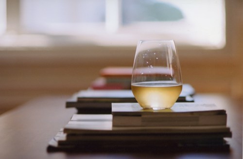 A glass of light-colored beverage sits on a stack of books, with soft natural light in the background.