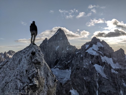 A person stands on a rocky peak, overlooking majestic snow-capped mountains under a cloudy sky.