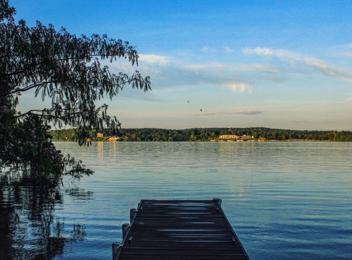 A serene lake view from a wooden dock, surrounded by trees and distant hills under a clear blue sky.