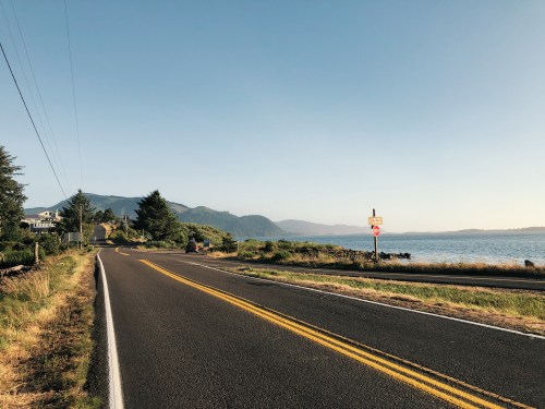 A scenic road curves along a waterfront, with mountains in the background and a clear blue sky above.