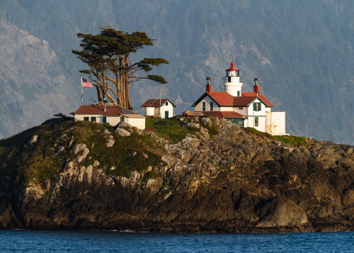 A scenic lighthouse on a rocky island, surrounded by trees and water, with an American flag waving in the breeze.