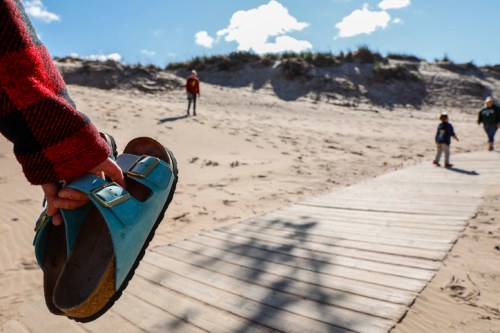 A person holds a blue sandal while walking on a sandy beach, with two children in the background.