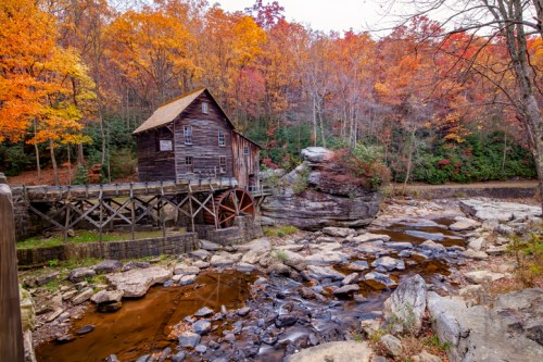 A rustic wooden mill beside a rocky stream, surrounded by vibrant autumn foliage.