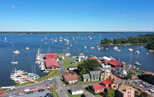 Aerial view of a marina with boats, a lighthouse, and colorful buildings along the waterfront under a clear blue sky.