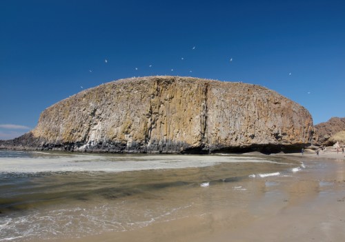 A large, rocky outcrop rises from a sandy beach under a clear blue sky, with seagulls flying above.