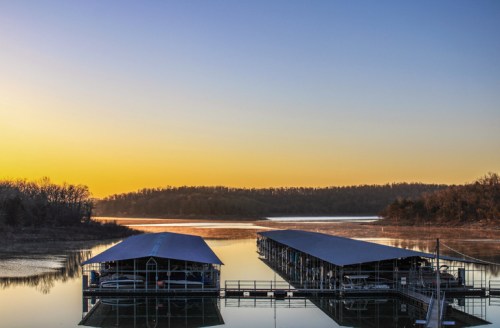 Sunrise over a calm lake with two boat docks and trees in the background, reflecting warm colors on the water.