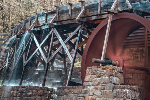 A wooden water wheel turns beside a stone structure, with water cascading down in a serene, natural setting.