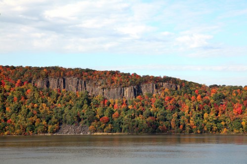 Vibrant autumn foliage on a rocky cliff overlooking a calm river under a partly cloudy sky.