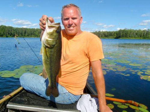 A man in an orange shirt holds a large bass fish while sitting on a boat in a lake surrounded by greenery.