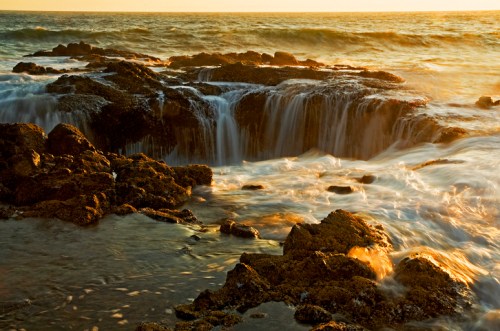 Sunset over rocky shoreline with gentle waves cascading over the rocks, creating a serene coastal scene.