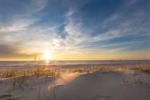 Sunset over a tranquil beach, with gentle waves and grass-covered sand dunes in the foreground.