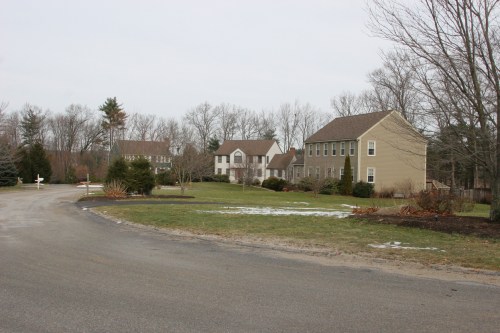 A quiet suburban street with several houses, bare trees, and patches of snow on the ground.