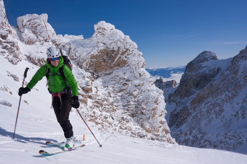 A skier in a green jacket ascends a snowy mountain slope with rocky peaks in the background under a clear blue sky.