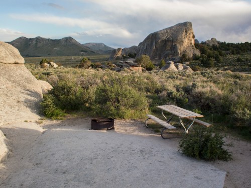 A picnic table and fire pit in a rocky, grassy landscape with mountains in the background under a cloudy sky.