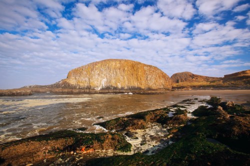 A rocky coastline with a large cliff, tide pools, and a cloudy blue sky in the background.