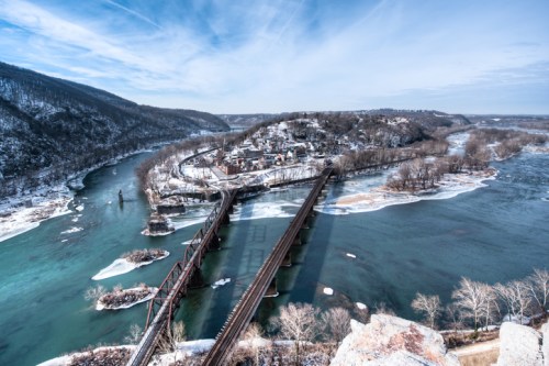 A scenic winter view of a river, a town, and a bridge surrounded by snow-covered hills and icy waters.