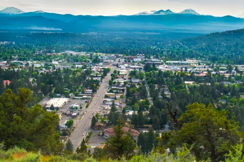 Aerial view of a small town surrounded by mountains, with a main road running through and greenery in the foreground.