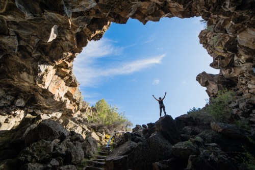 A person stands triumphantly in a rocky cave, arms raised, with a bright blue sky visible through the opening above.