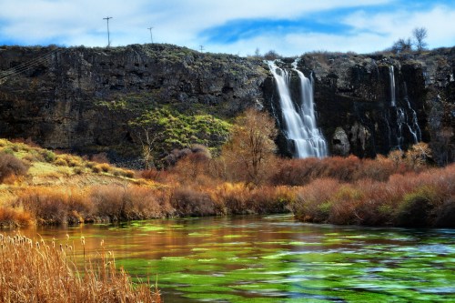 A scenic view of a waterfall cascading down a rocky cliff, surrounded by autumn foliage and a reflective green river.