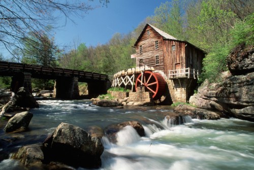 A historic wooden mill with a large water wheel beside a flowing river, surrounded by lush greenery and a blue sky.